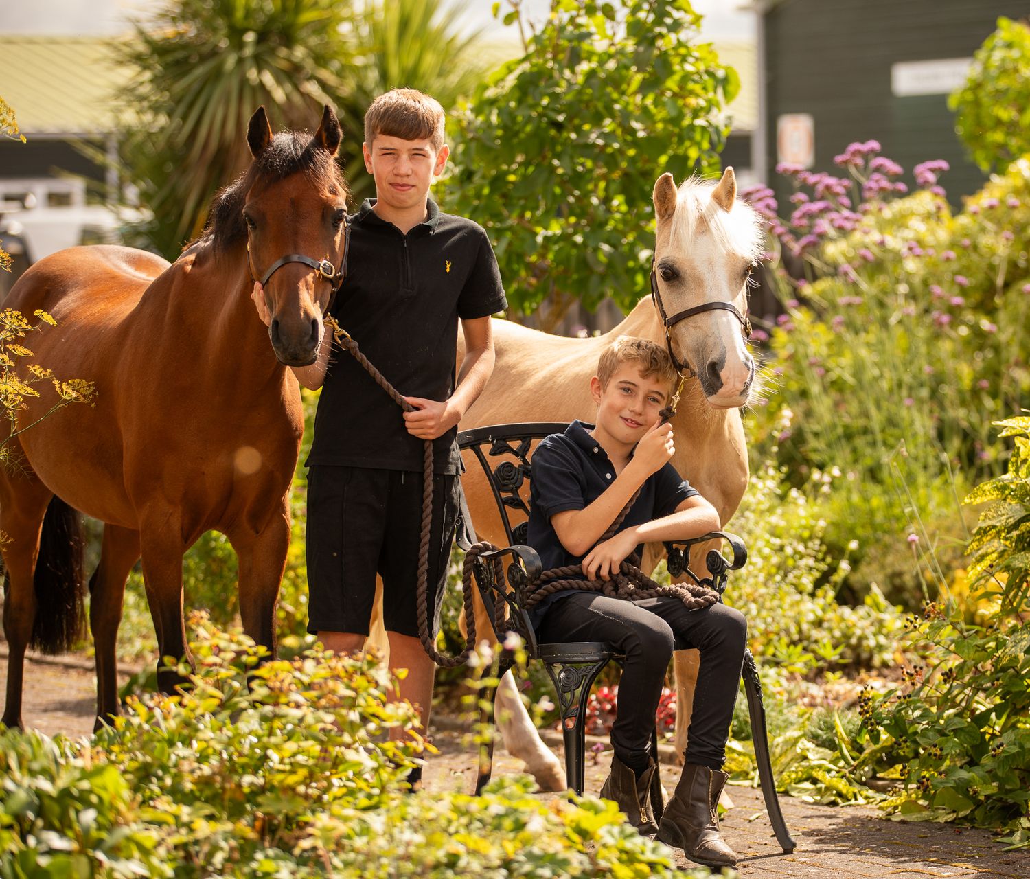 Two horses stand with people in a lush garden setting on a sunny day with colorful flowers blooming.