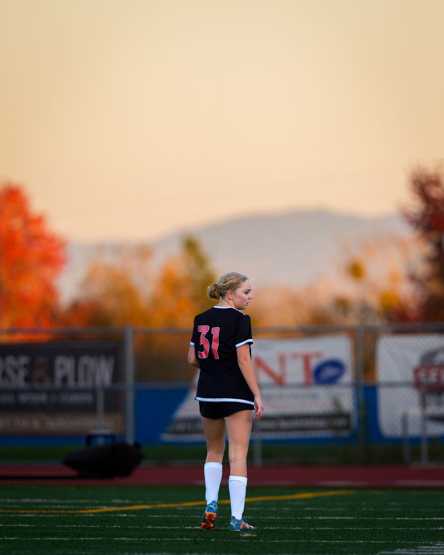 A soccer player kicks the ball on a turf field at sunset with autumn trees in the background.