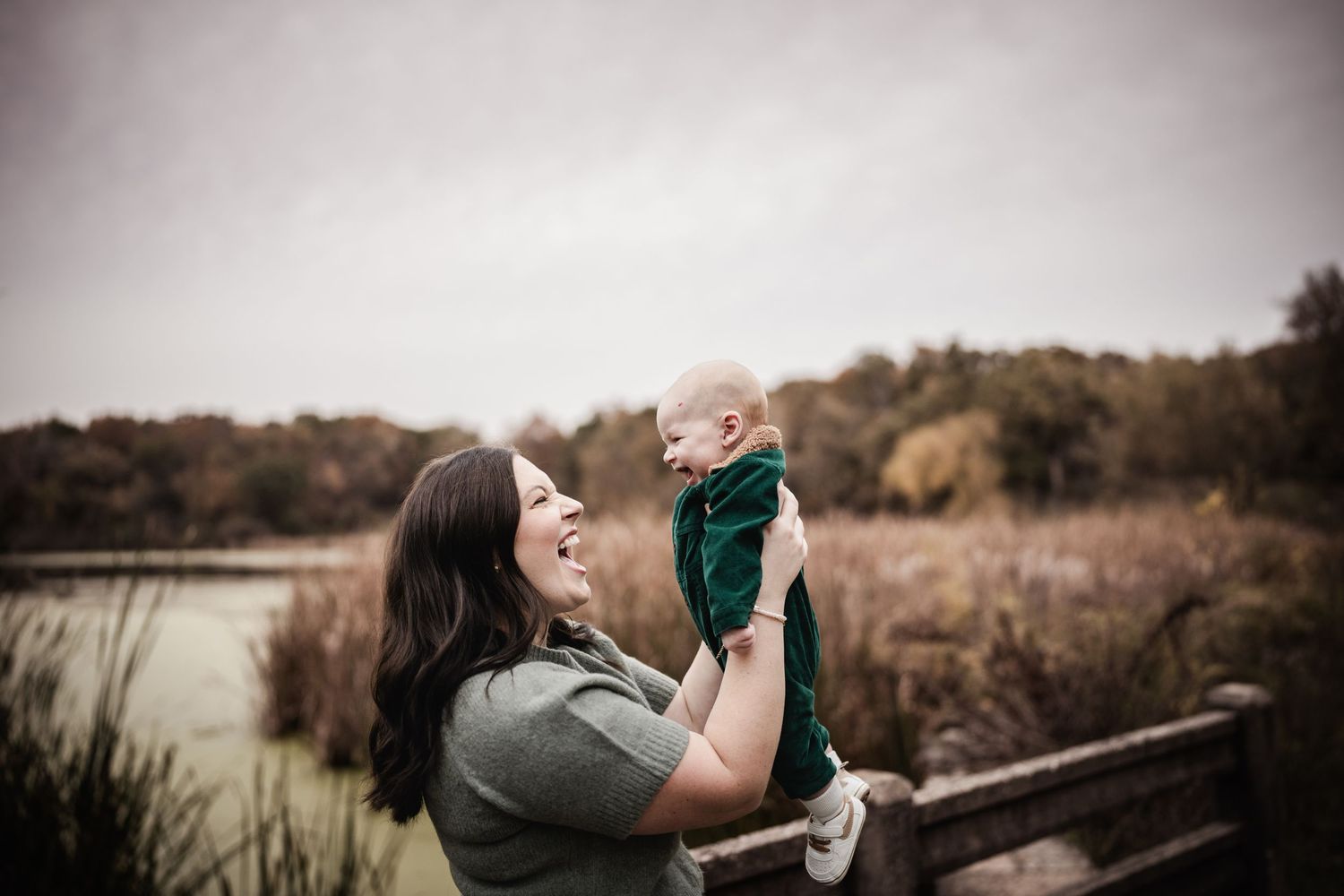 A parent lifts a small child wearing green outdoors near a pond on an autumn afternoon during a photo session in Palatine