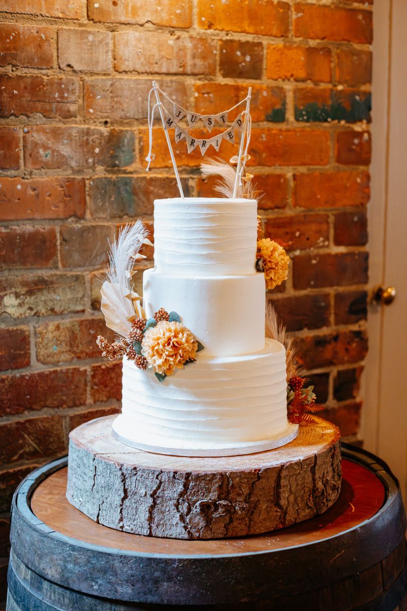 Three-tiered white wedding cake with rustic wood slice base and floral accents displayed against exposed brick wall.