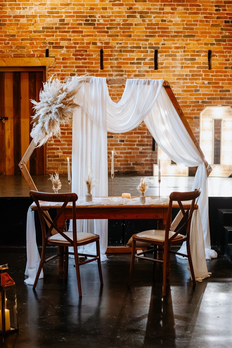 Elegant white fabric draping and wooden chairs create a romantic atmosphere against exposed brick walls in a rustic venue.