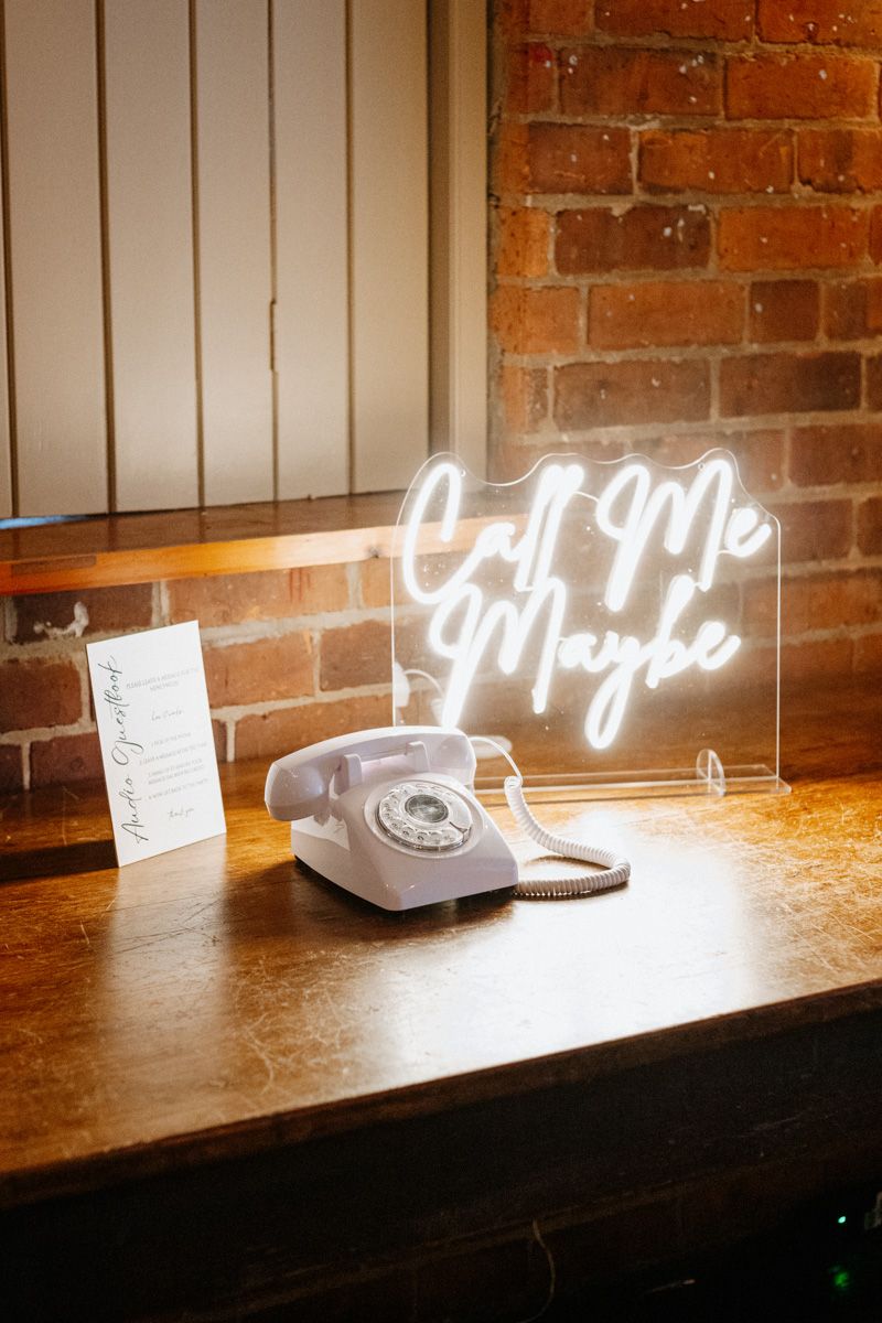 Vintage rotary telephone and neon sign display on wooden table against brick wall with dramatic lighting.