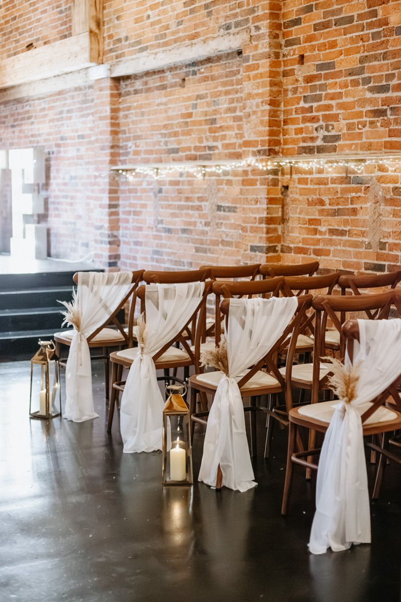 Rustic wedding venue with wooden chairs draped in white fabric against exposed brick walls.
