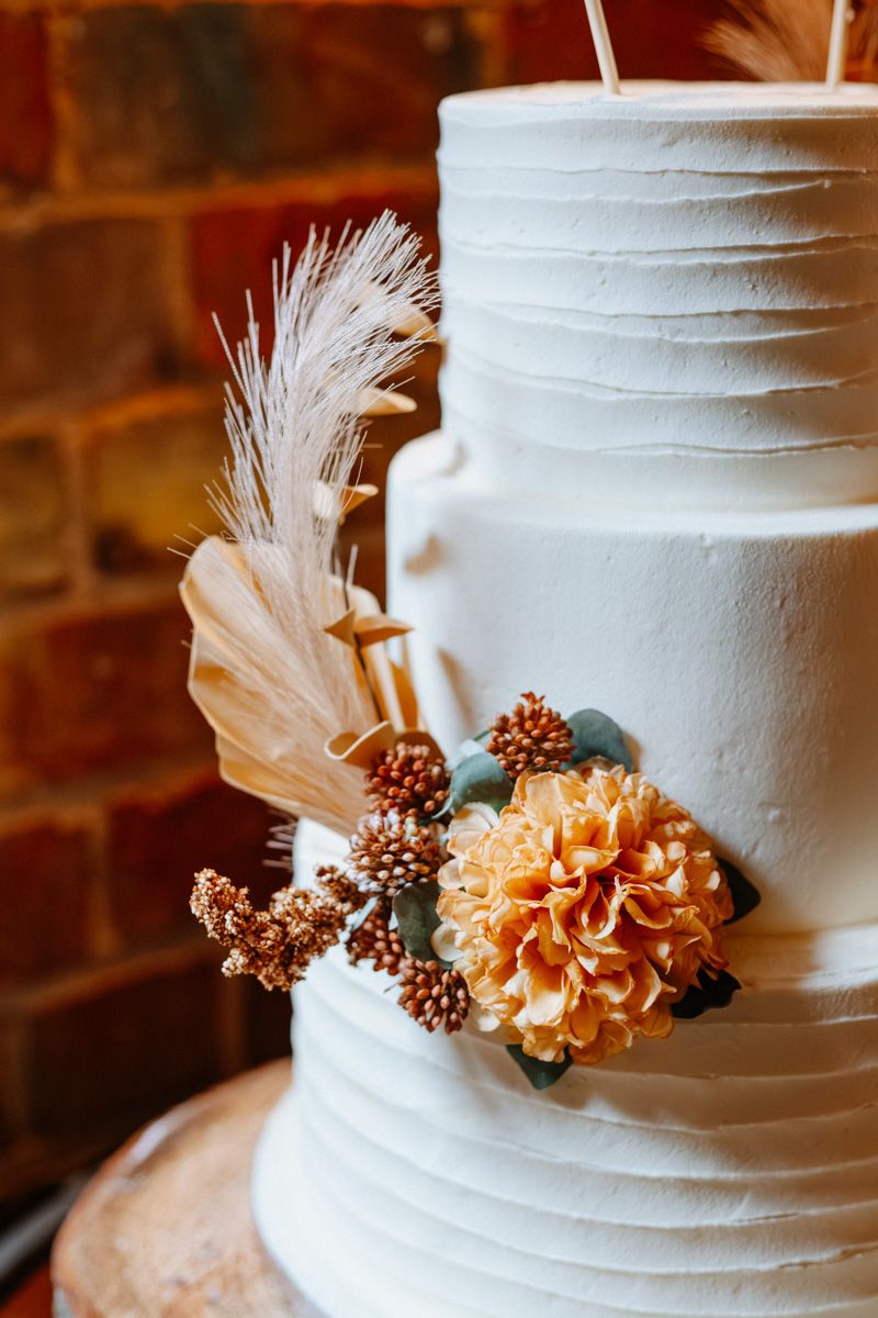 White wedding cake decorated with delicate peach flowers, pinecones, and feathers against a rustic brick wall background.