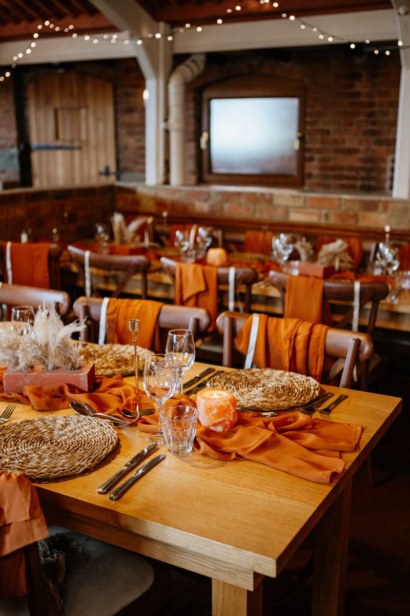 Wedding reception tables set with orange linens and woven chargers in an industrial brick-walled venue.
