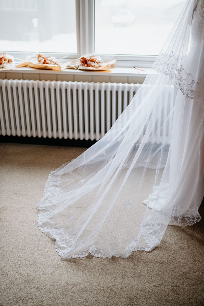 Close-up view of a white lace wedding dress hem trailing along beige carpet near a window.