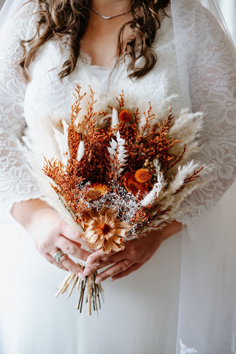 Dried orange and white floral wedding bouquet held against a white lace wedding dress.