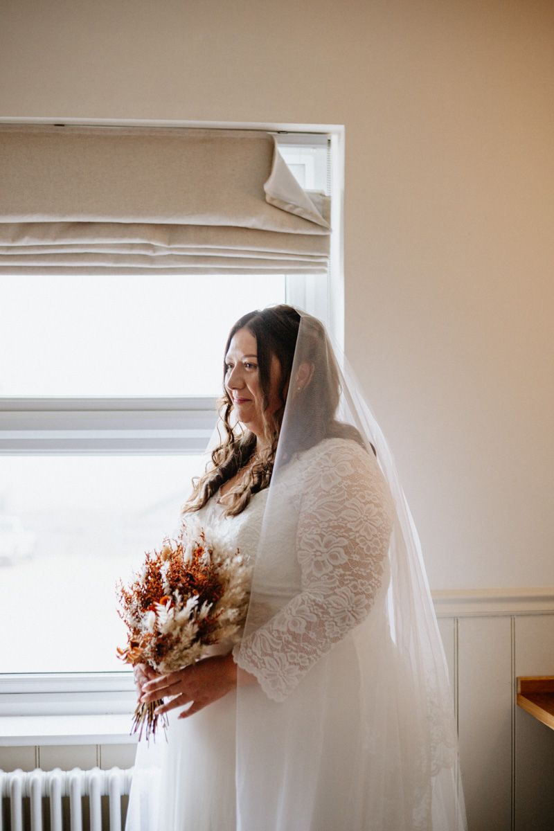 A bride in a white lace wedding dress holds a dried autumn bouquet while standing by a window in soft natural light.