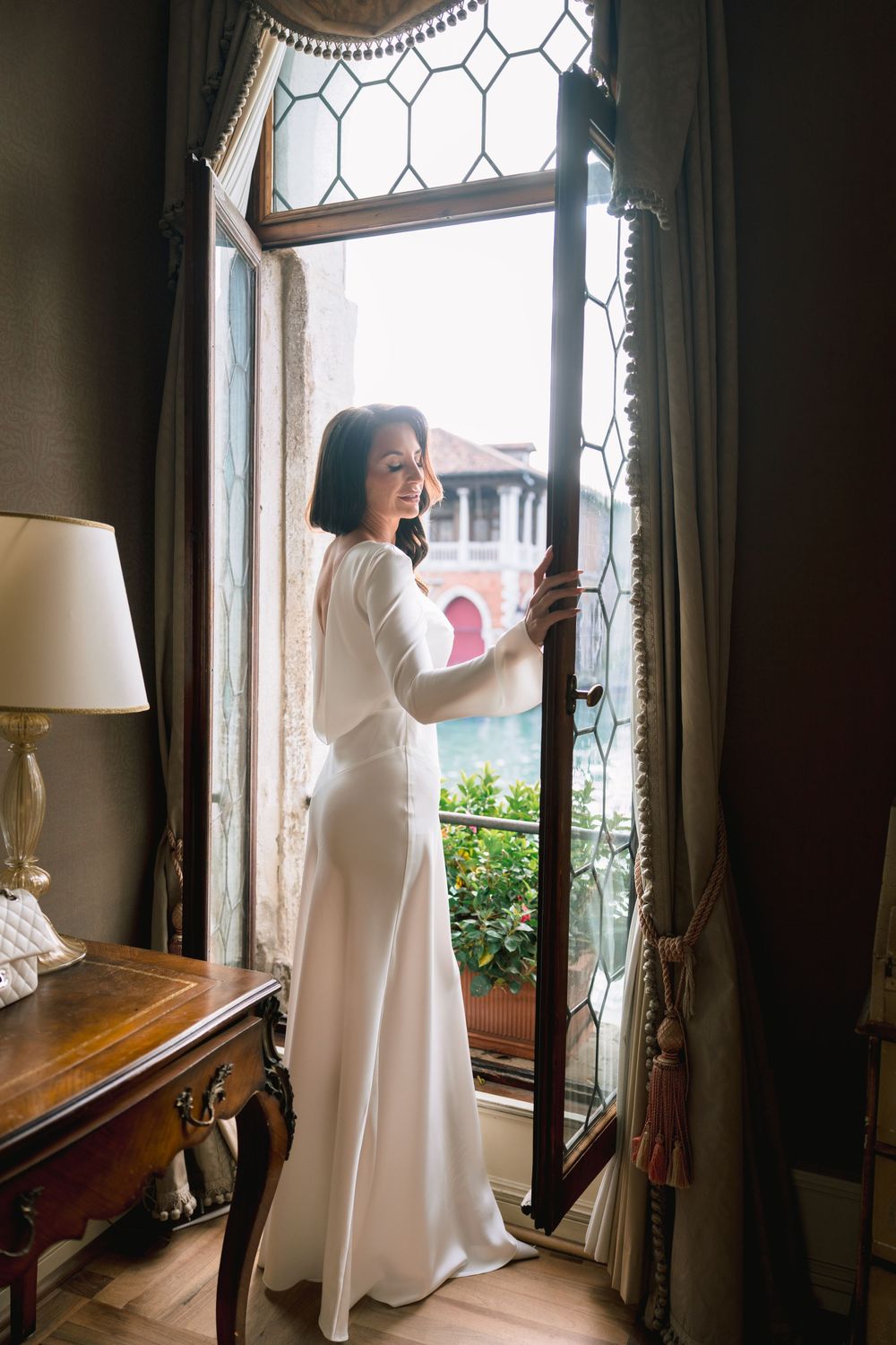A person in a flowing white dress stands by an ornate window with leaded glass panes in a vintage interior.