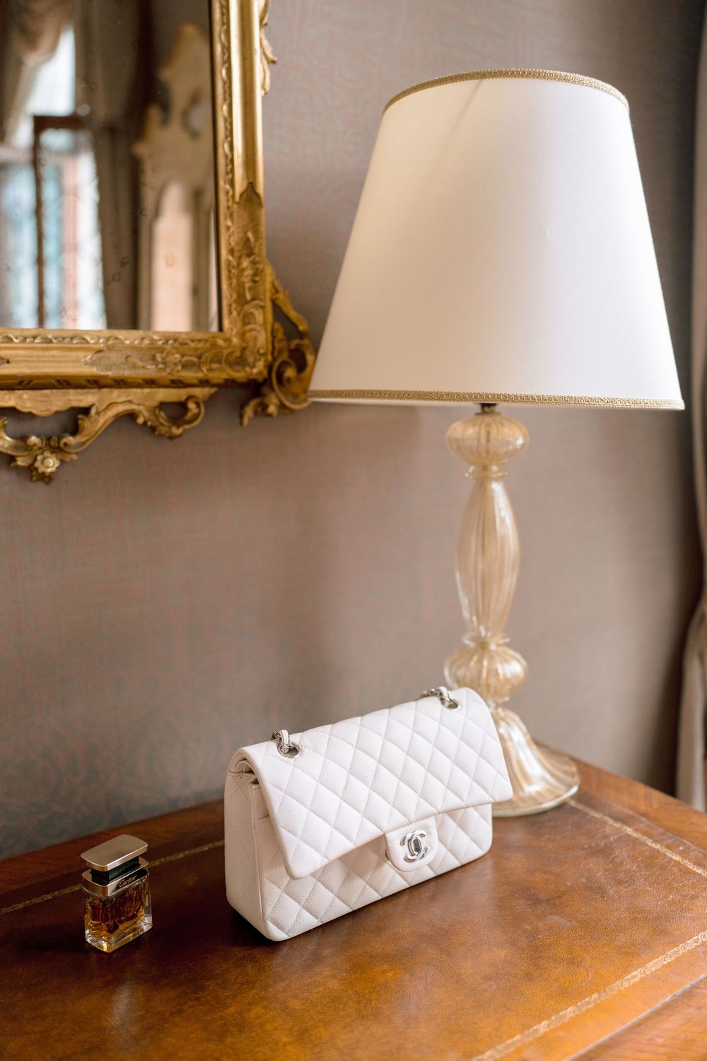 A white quilted Chanel handbag sits elegantly on a wooden table next to a crystal table lamp with white shade.