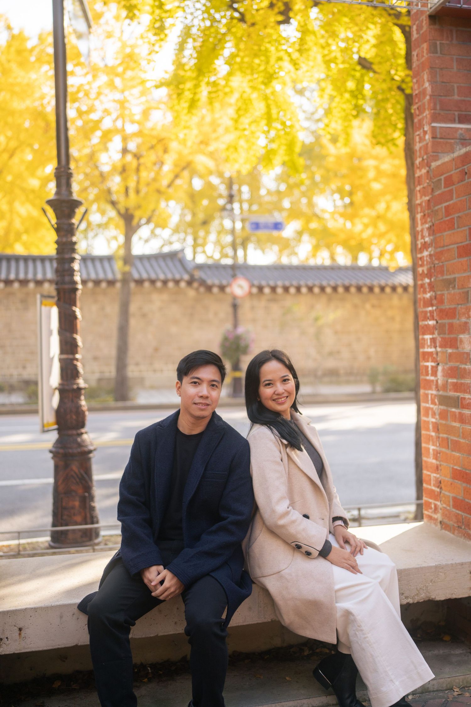 A couple sits together on a brick ledge with autumn trees creating a warm golden backdrop.
