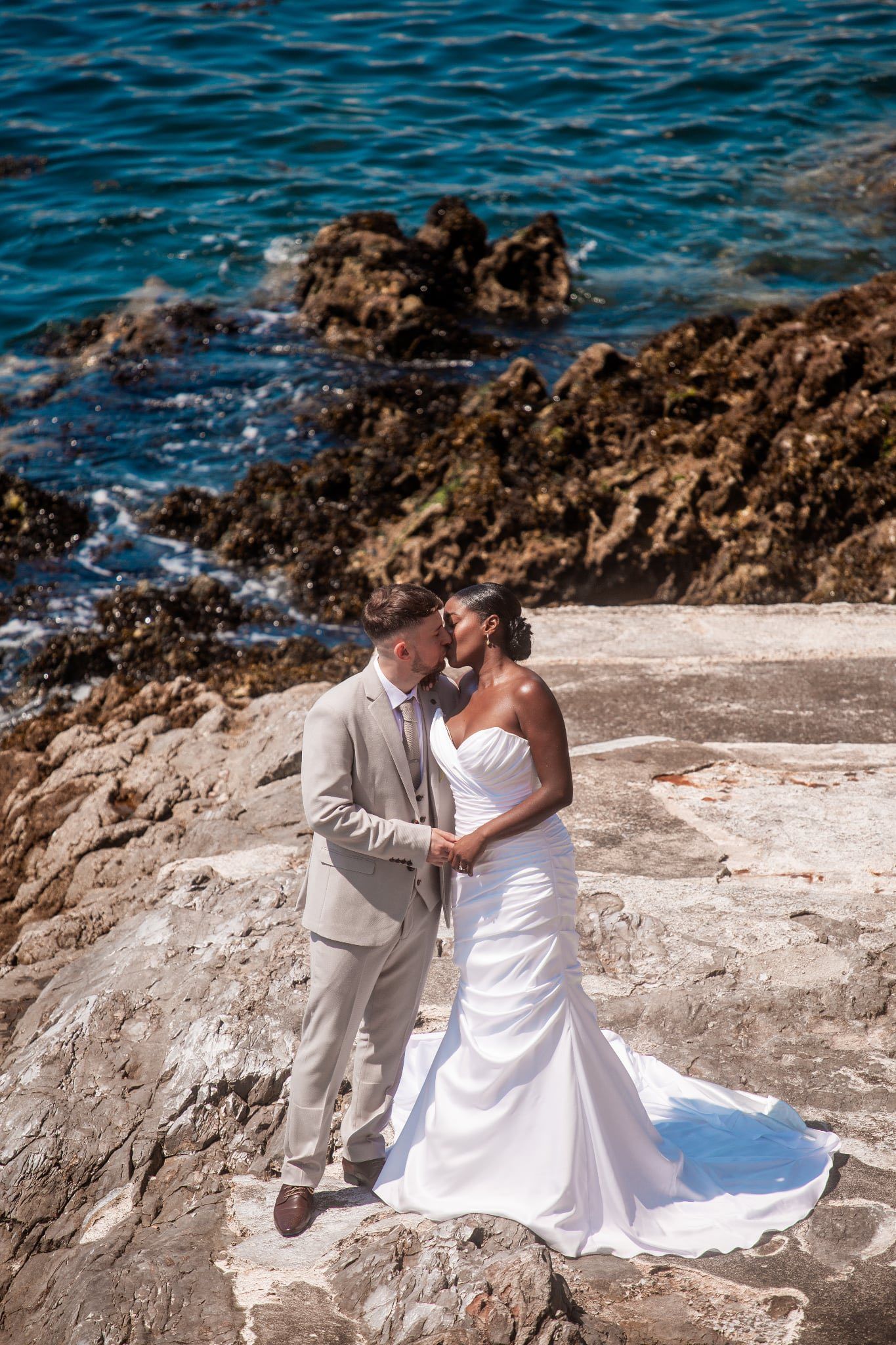 A couple in wedding attire poses on rocky coastal cliffs near blue ocean waters. Pier One wedding, Plymouth