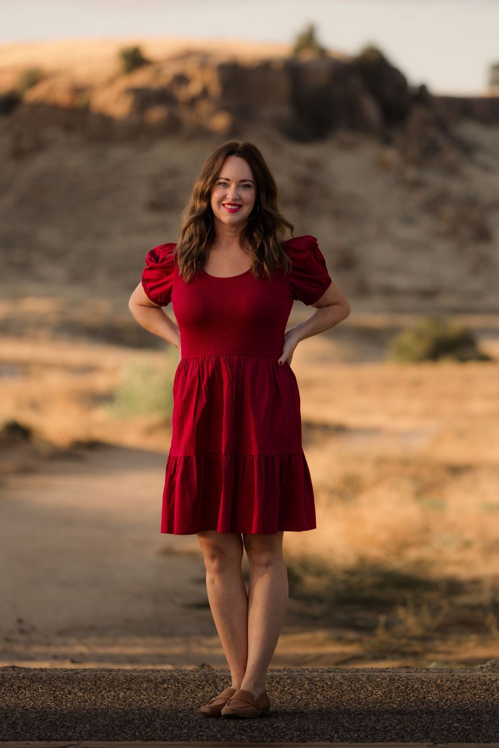 Woman in flowing red dress poses confidently in golden desert landscape.