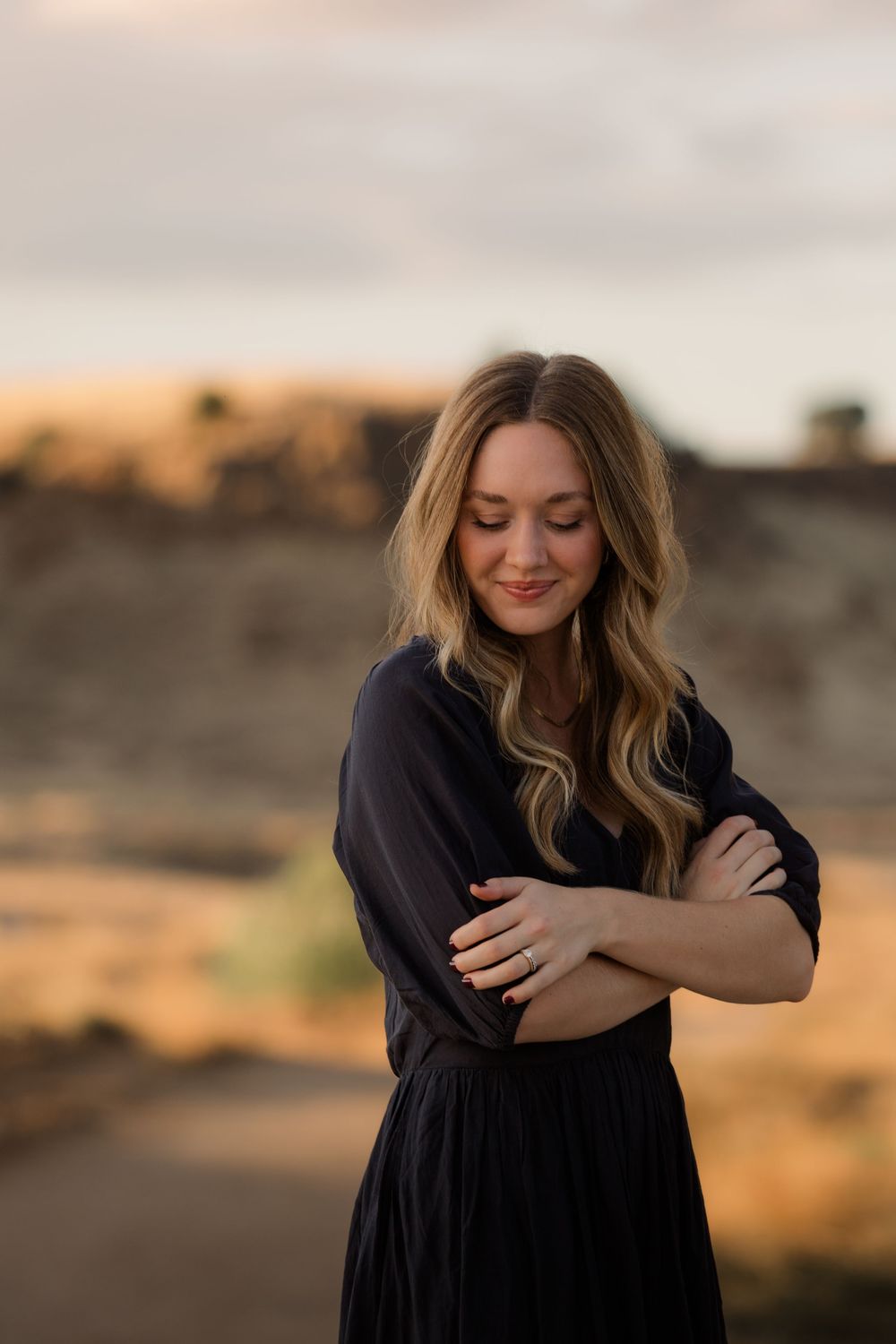 Woman in black dress hugs herself while standing in golden sunset light.
