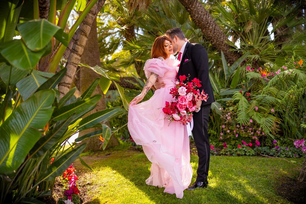 A couple in formal attire embraces in a lush tropical garden setting with vibrant greenery and pink flowers.