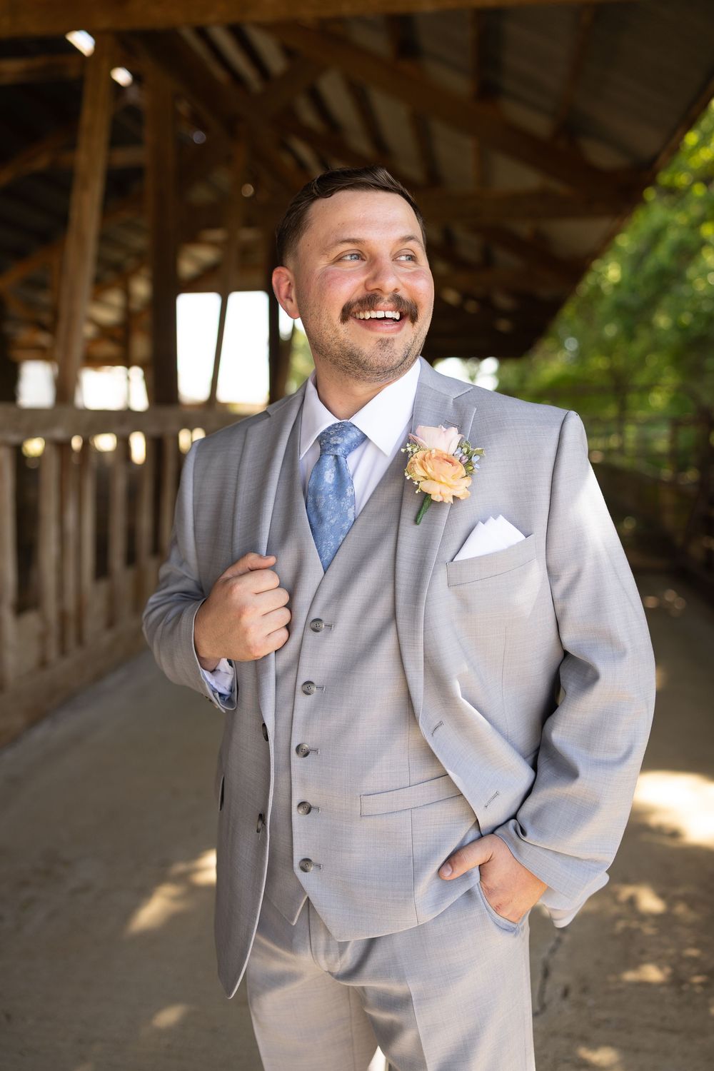 Groom in light gray three-piece suit with blue tie and floral boutonniere smiling at outdoor wedding venue with rustic barn.