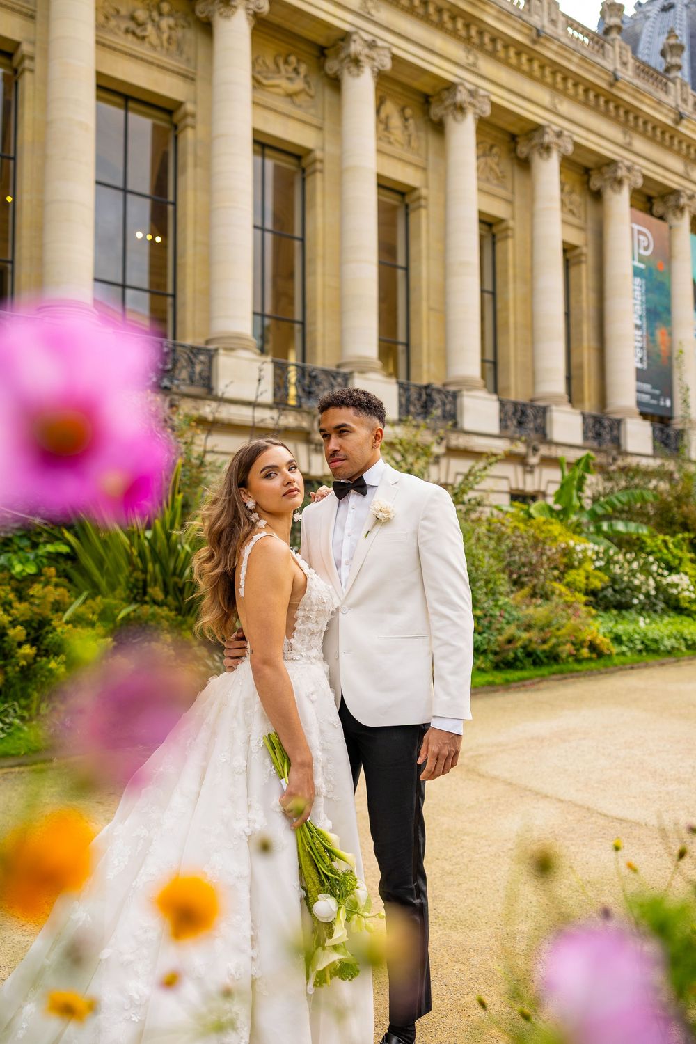 Elegant couple in formal attire poses together against ornate classical architecture with colorful flowers in foreground.
