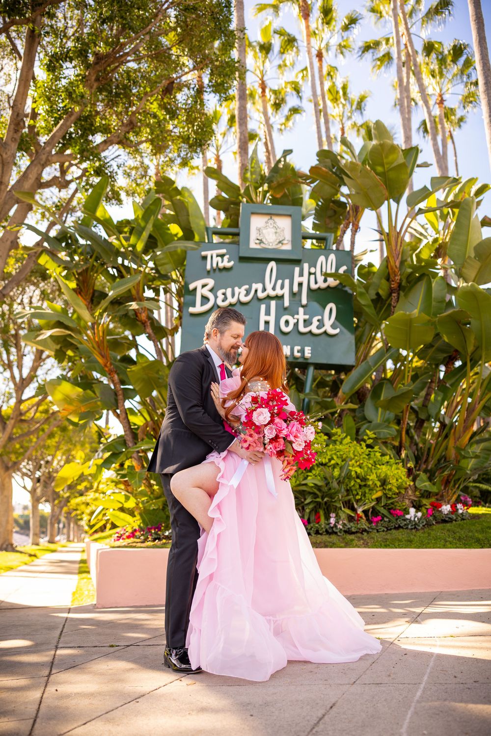 A couple shares a romantic moment on a pink bench outside the Beverly Hills Hotel surrounded by lush palm trees.