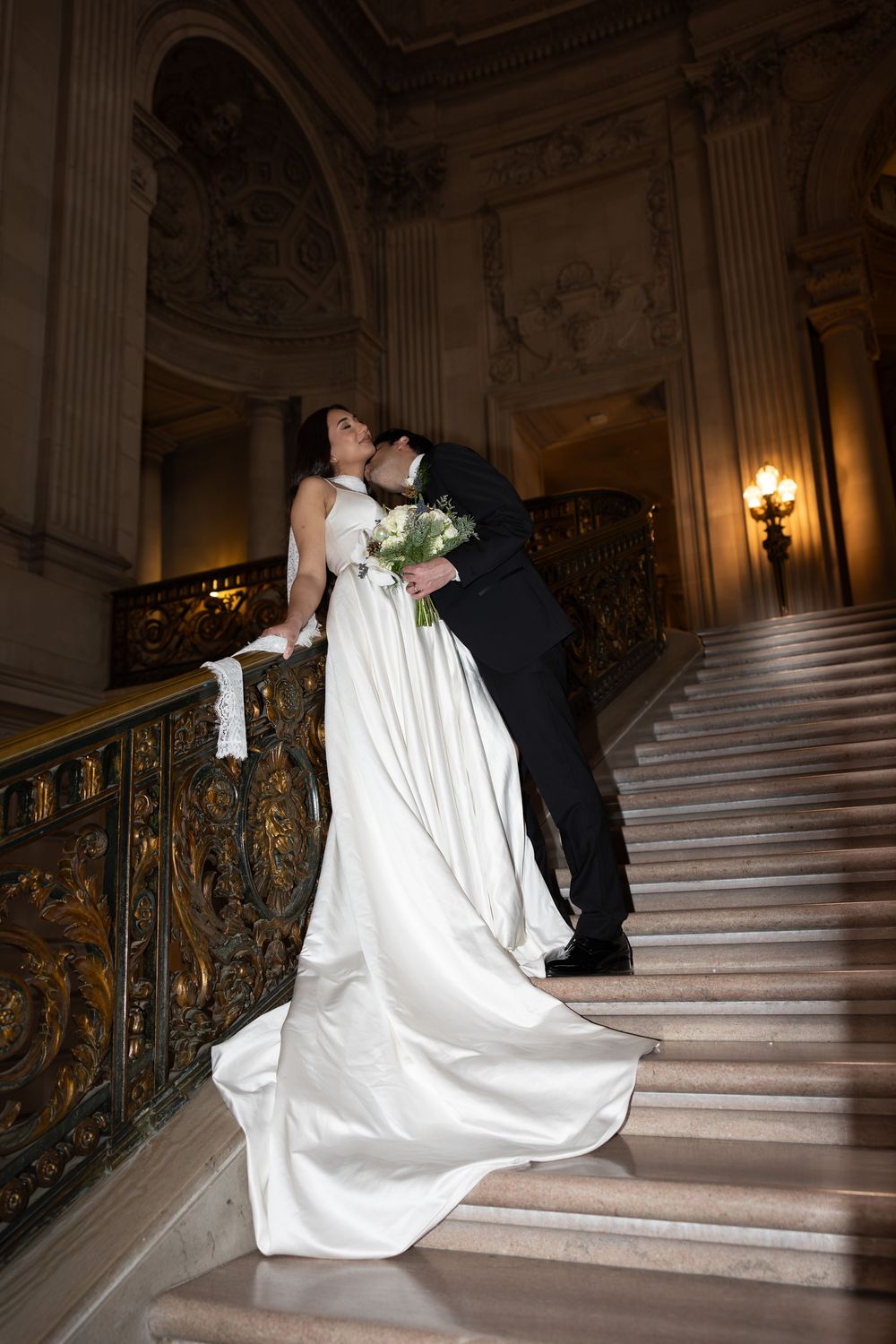 A bride and groom share a romantic moment on an elegant marble staircase with ornate gold railings.