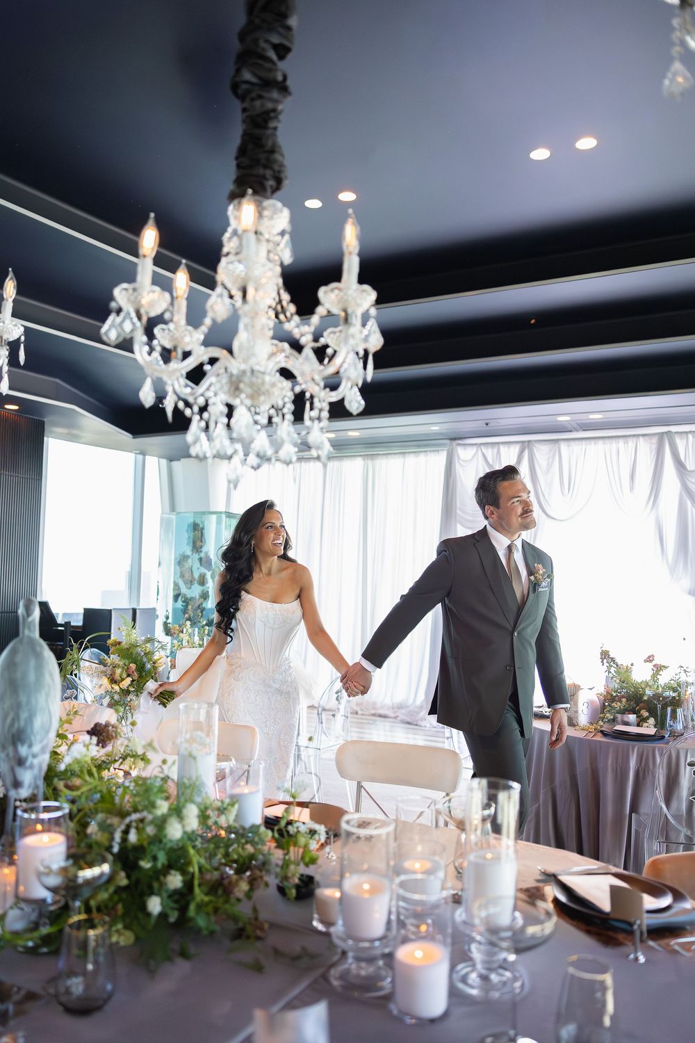 Bride and groom hold hands at elegantly decorated wedding reception table with crystal chandelier and candles.