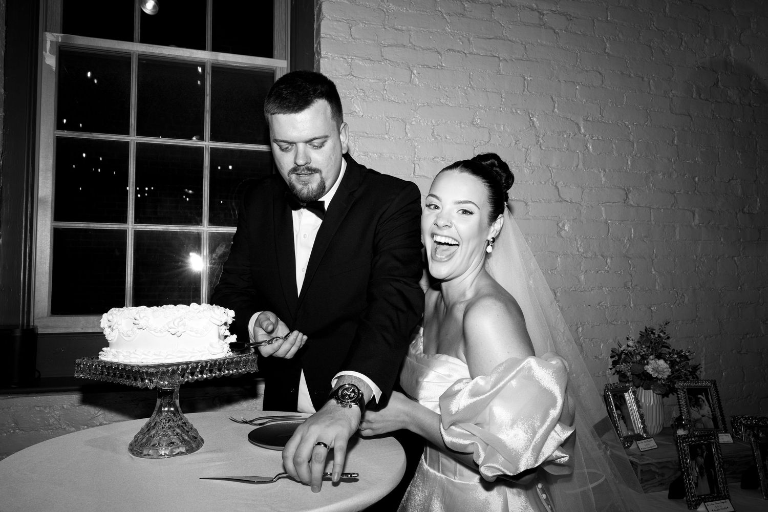 A bride in a white gown makes a heart shape with her hands while a man in a tuxedo waves at a nighttime celebration.
