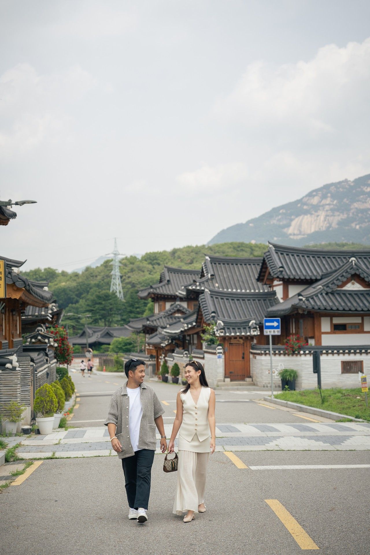 A couple walks hand in hand down a street in a traditional Korean neighborhood with mountains in the background.