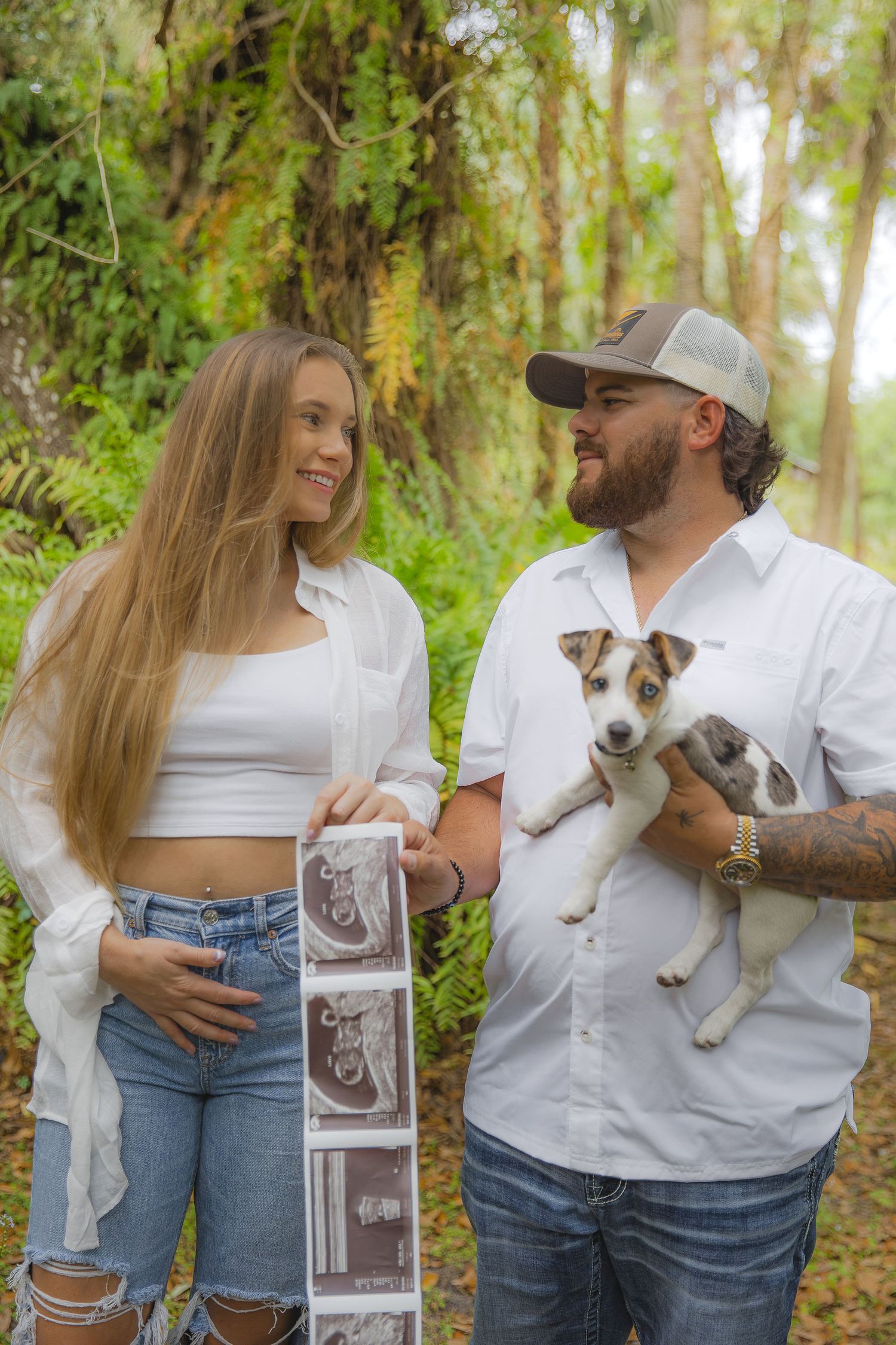 A couple holding a small dog poses together in a wooded area while displaying ultrasound photos.