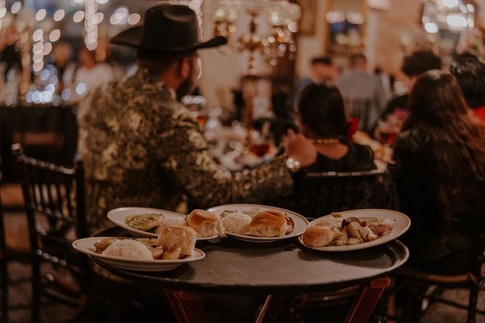 Plates of food served on round table in dimly lit restaurant with person wearing hat.