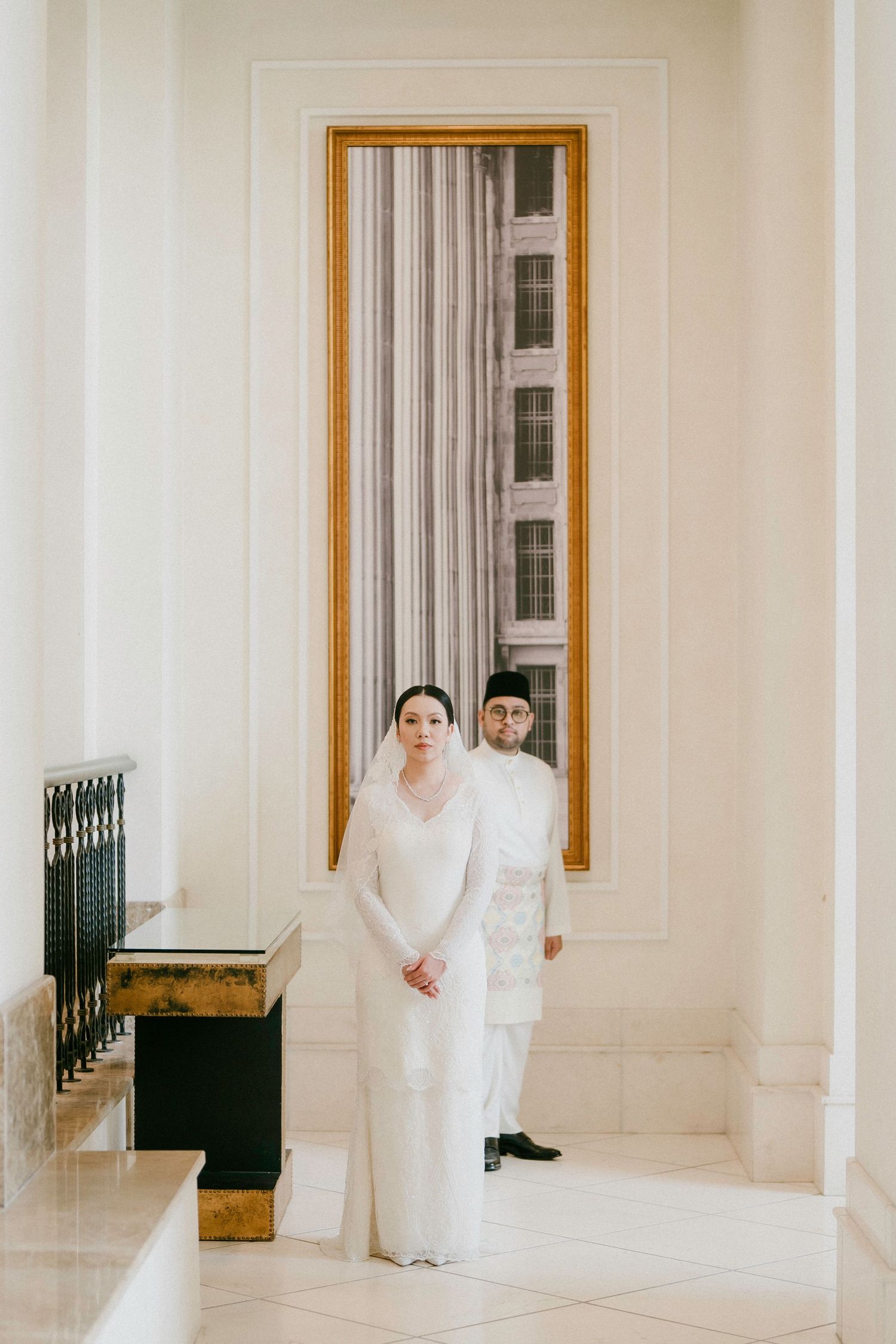 Elegant couple in white traditional Malay wedding attire standing in a grand white marble hall with ornate columns.