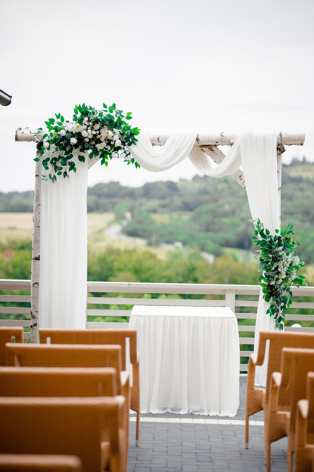 White wedding arch decorated with green foliage stands on a deck overlooking scenic hills with wooden chairs set up.