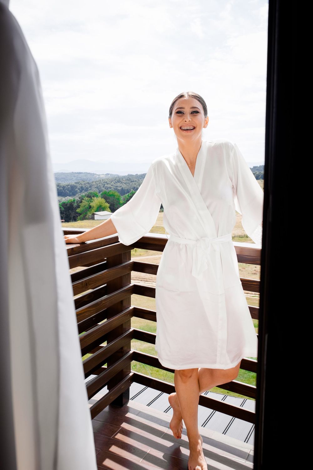 A person in a white robe stands on a wooden balcony overlooking a scenic mountain landscape on a sunny day.