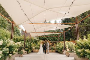 A couple stands under an elegant white canopy surrounded by lush greenery and hydrangeas during an outdoor ceremony.