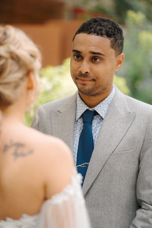 A groom in a light gray suit and navy blue tie smiles warmly during an outdoor wedding ceremony.