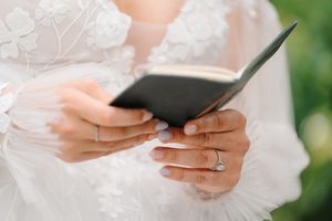 Close-up view of hands in white lace holding a small black book with a diamond ring on the finger.