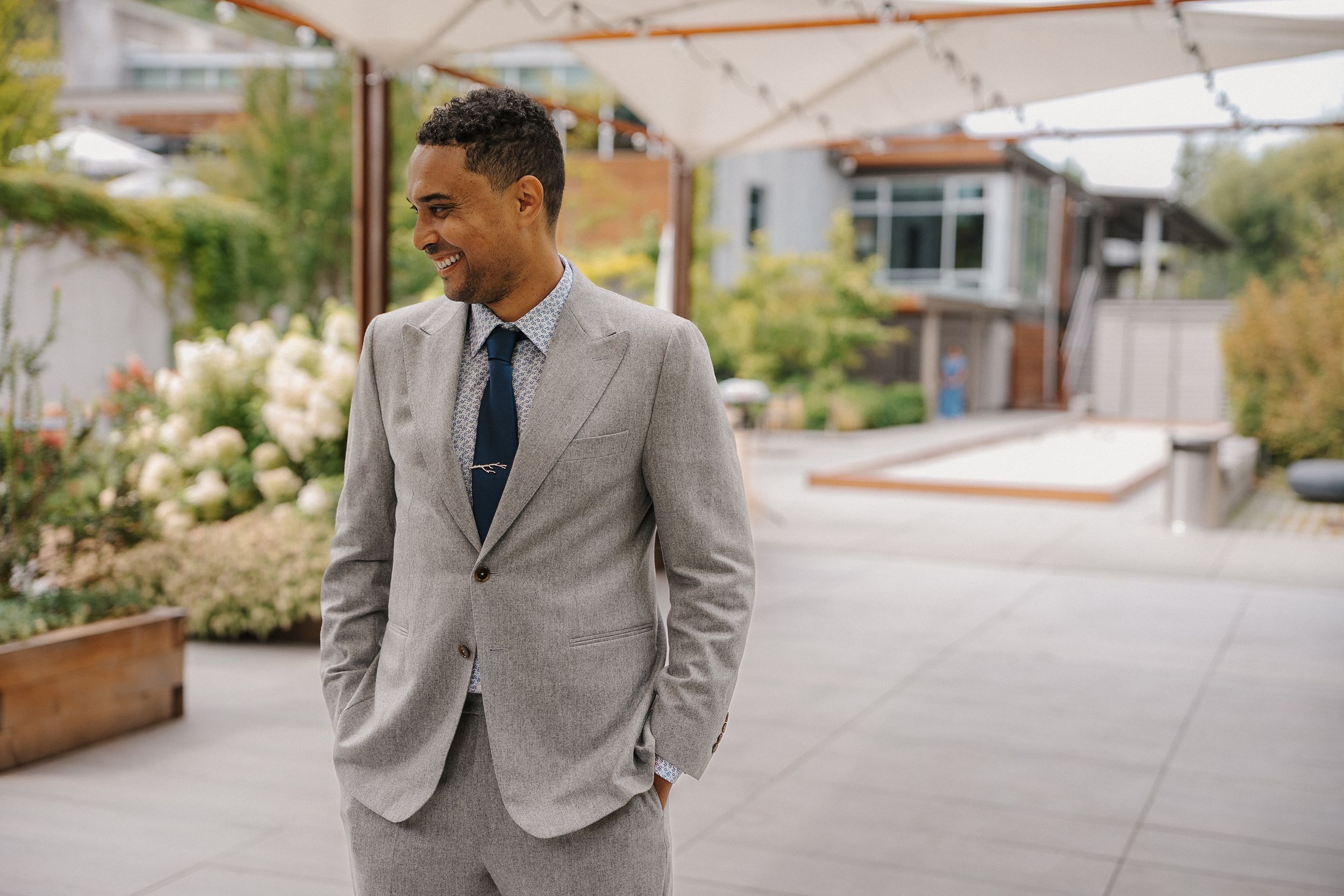 A person in a well-fitted gray suit stands confidently outdoors against a residential backdrop.