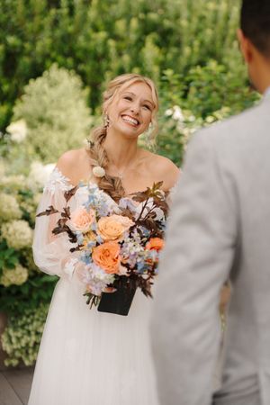 A bride in an off-shoulder wedding dress holds a peach and white floral bouquet while smiling outdoors.