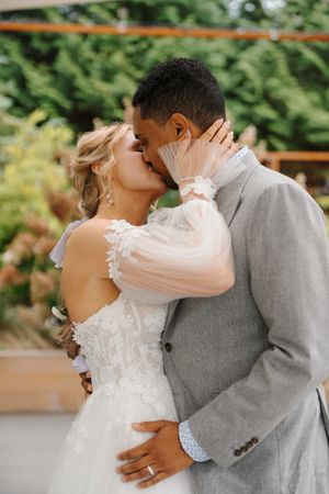 A romantic wedding couple shares an intimate kiss during their outdoor garden ceremony wearing white and grey attire.