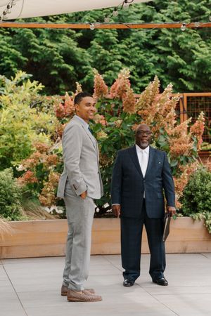 Two men in contrasting suits stand together in a garden setting with blooming astilbe flowers and planters.