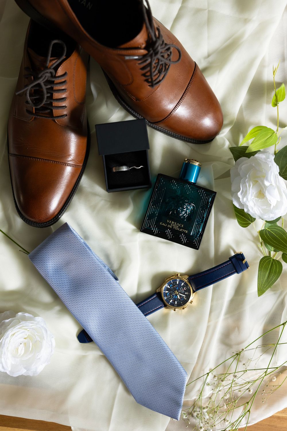 Wedding accessories including brown leather dress shoes, light blue tie, watch, and white flowers arranged on white fabric.
