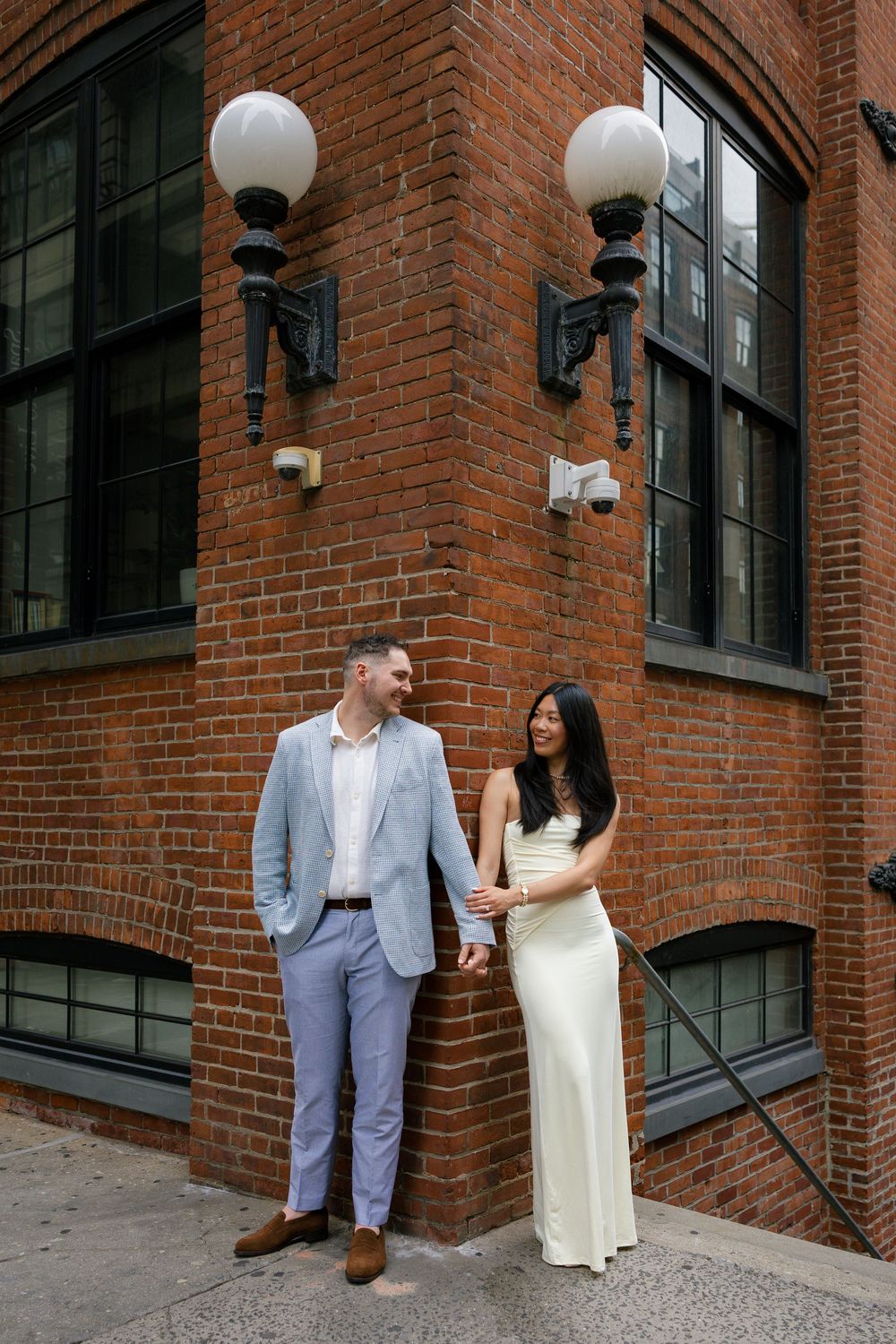 Couple holding hands smiling at each other in front of a brick building with globe lights.