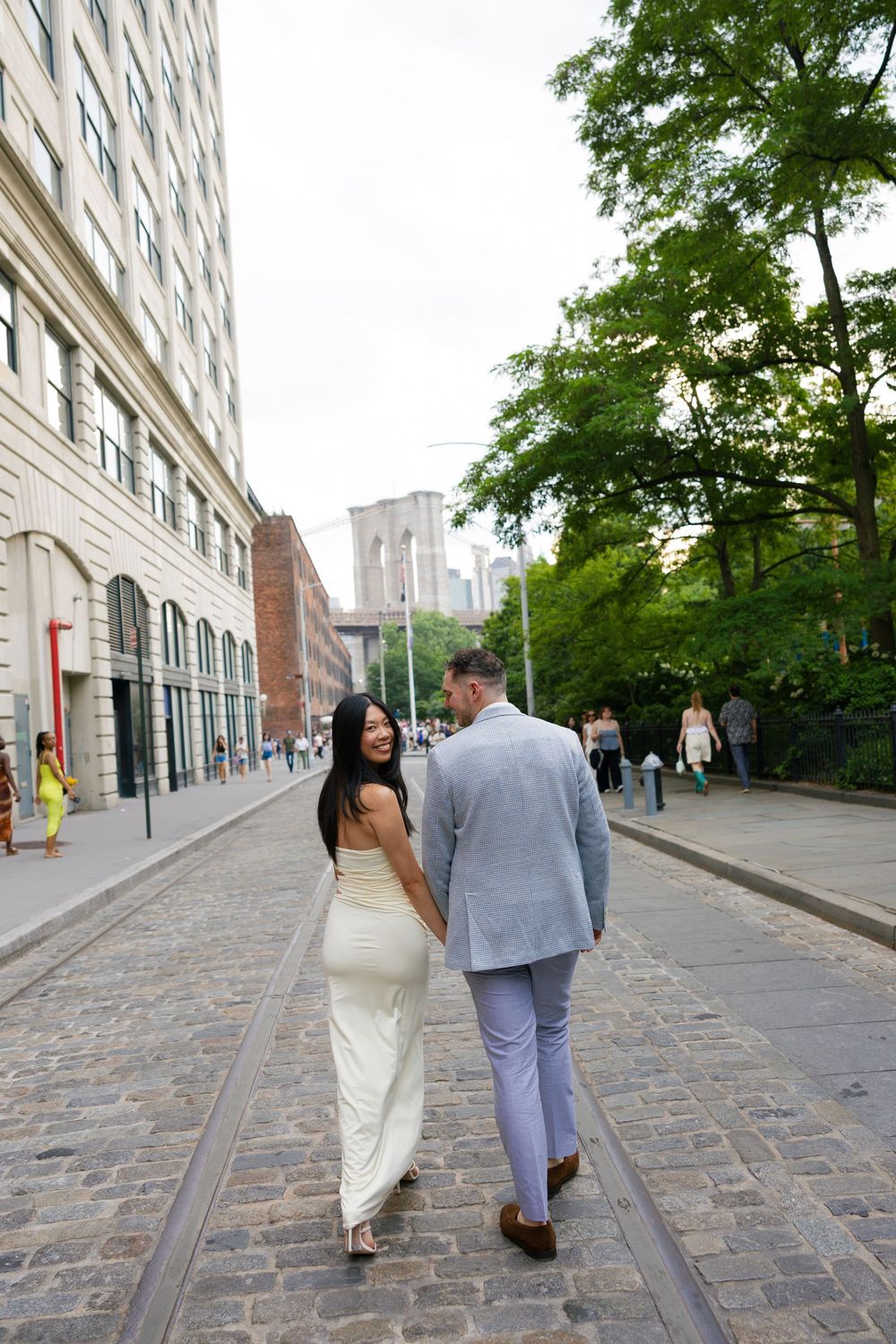 Couple walking hand-in-hand on cobblestone street near Brooklyn Bridge, woman in white dress smiling back.