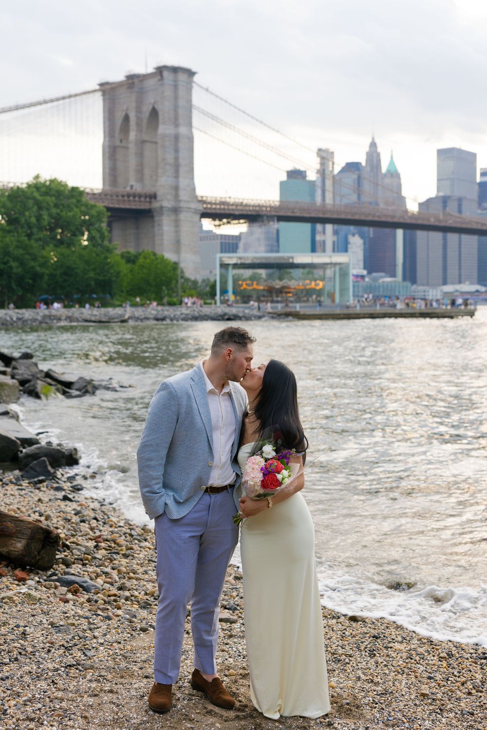 Couple kissing on rocky shore with Brooklyn Bridge and Manhattan skyline backdrop during wedding photo session.