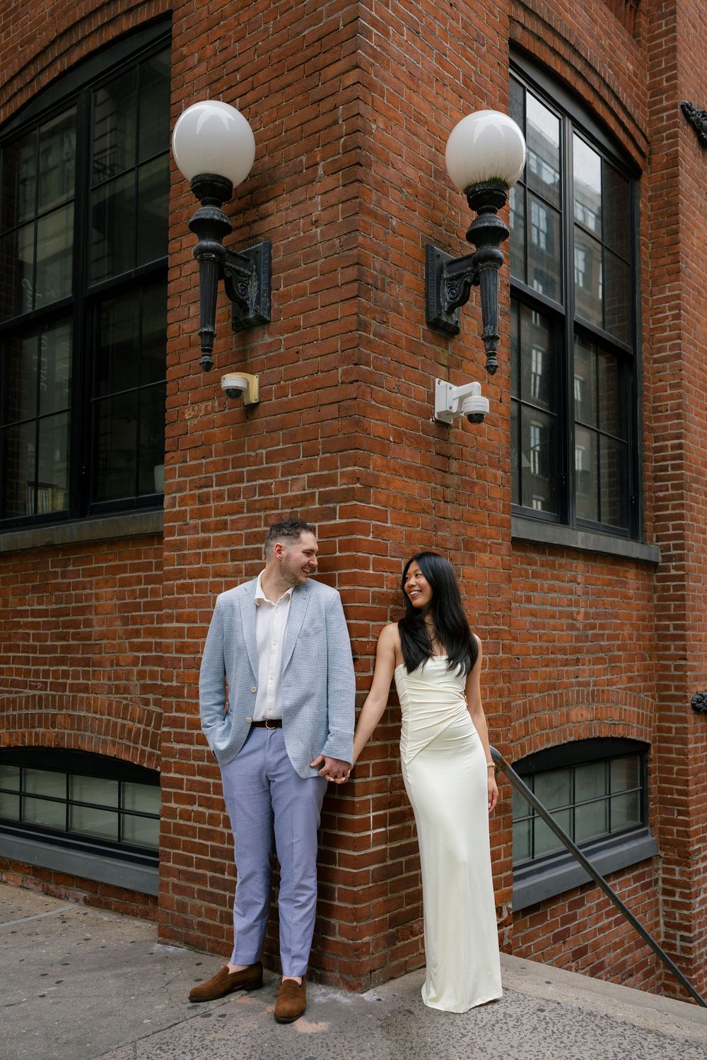 Couple in formal attire poses against a brick building with white globe lights in a series of romantic shots.