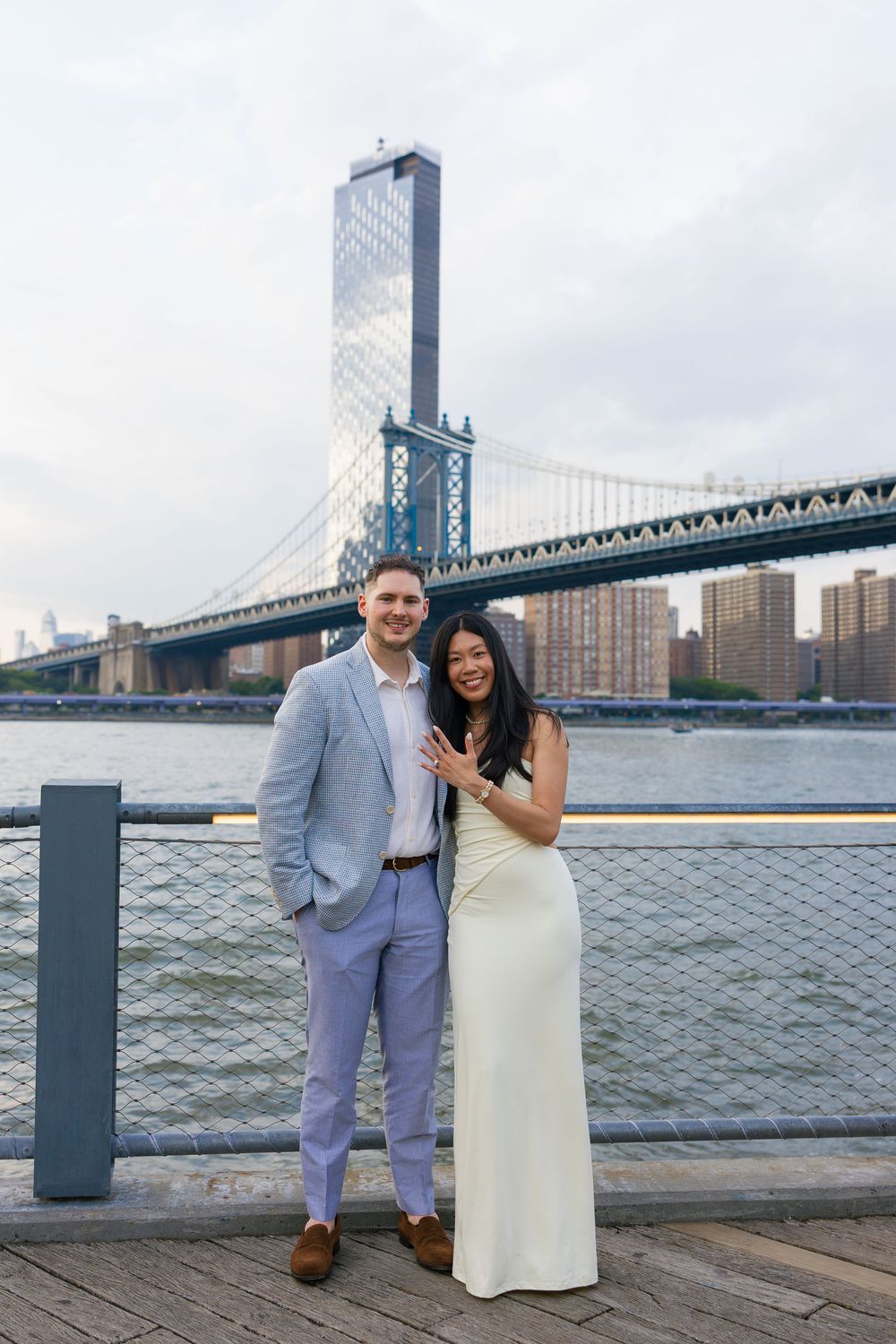 Couple posing by the Manhattan Bridge waterfront, woman in white gown showing engagement ring, man in blue suit.