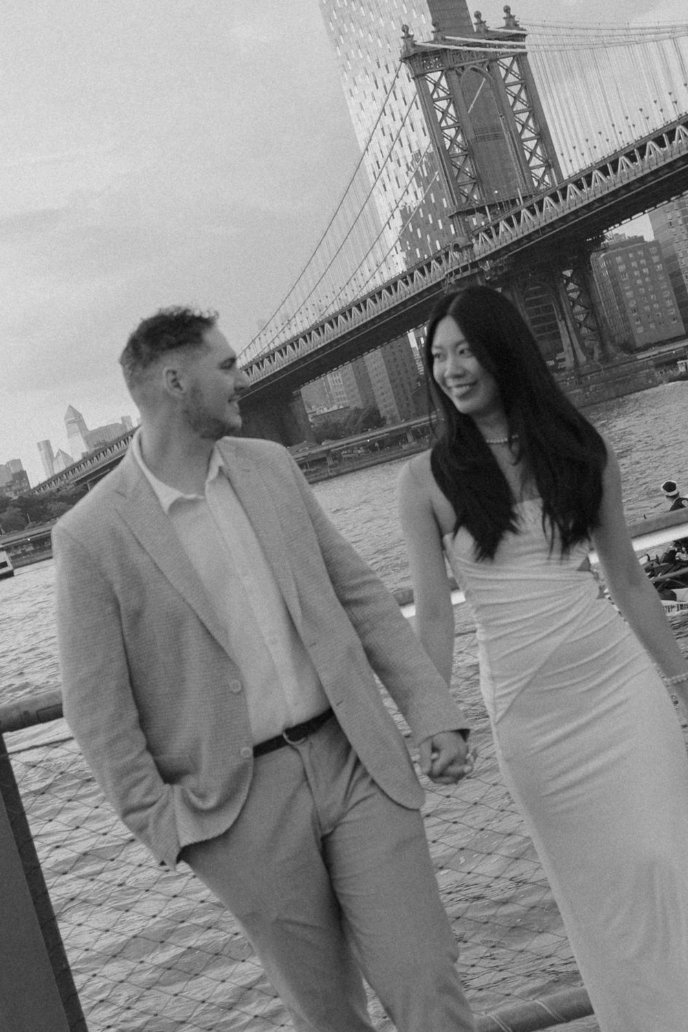 Black and white photo of a couple holding hands by the Manhattan Bridge waterfront in New York City.