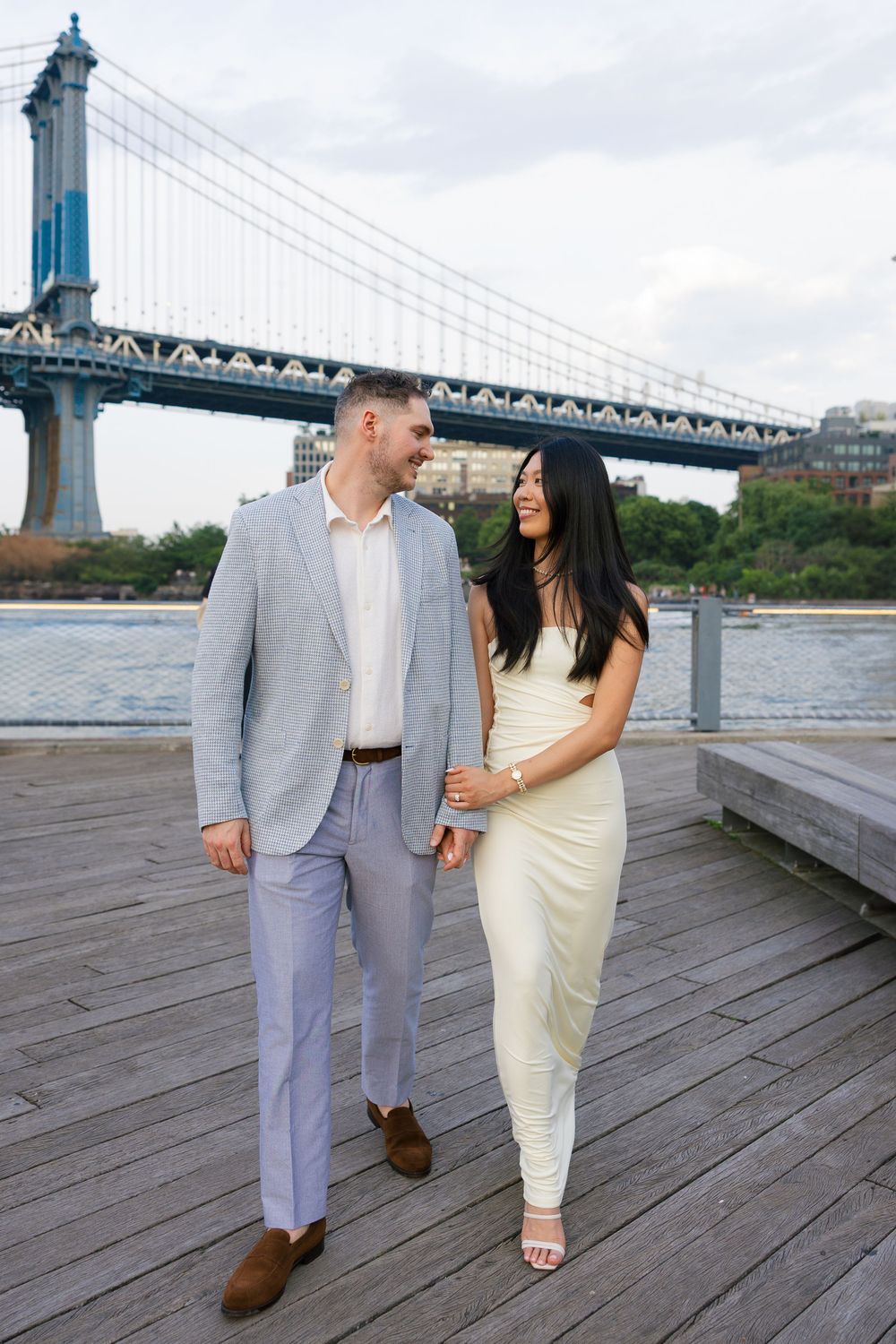 Couple walking on Brooklyn waterfront boardwalk with Manhattan Bridge backdrop, man in gray suit, woman in white dress.