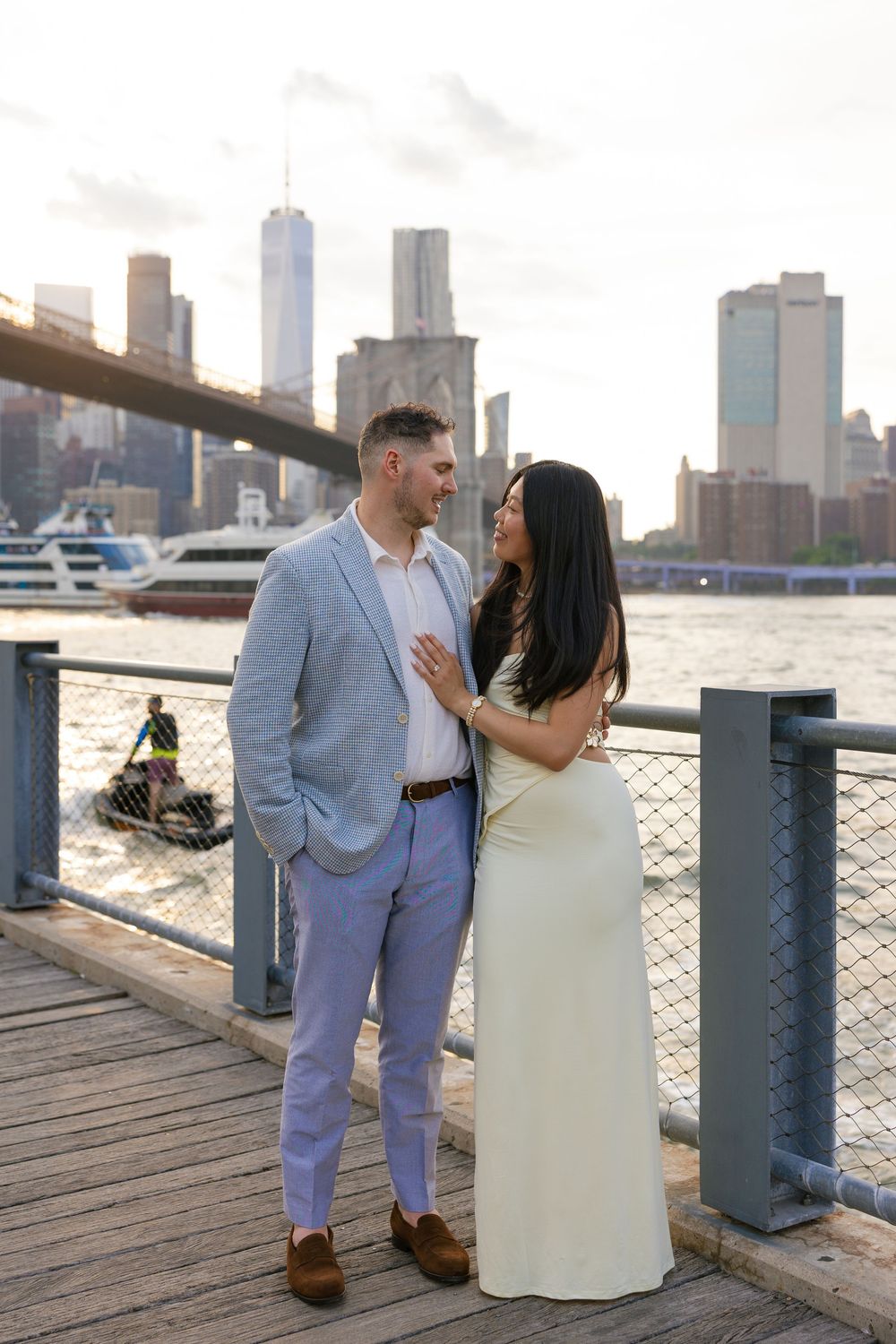Couple posing on Brooklyn Bridge Park waterfront pier with NYC skyline and Brooklyn Bridge at sunset.