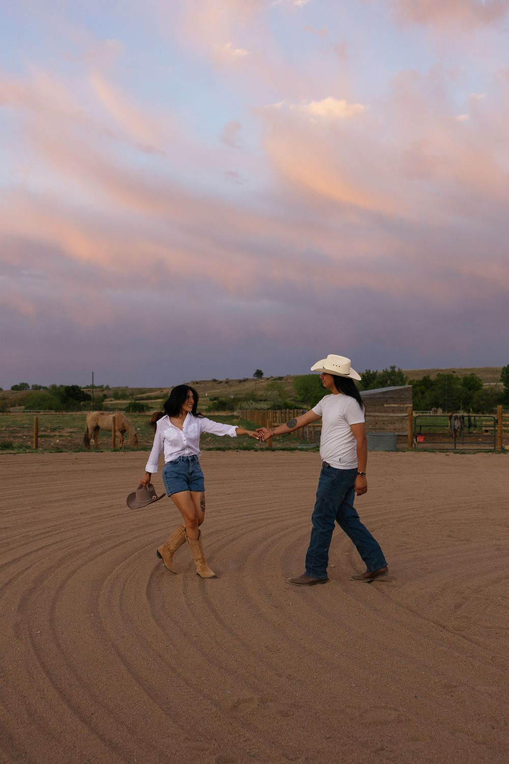 Couple holding hands dancing in a sandy ranch arena at sunset with horses and pink clouds in the background.
