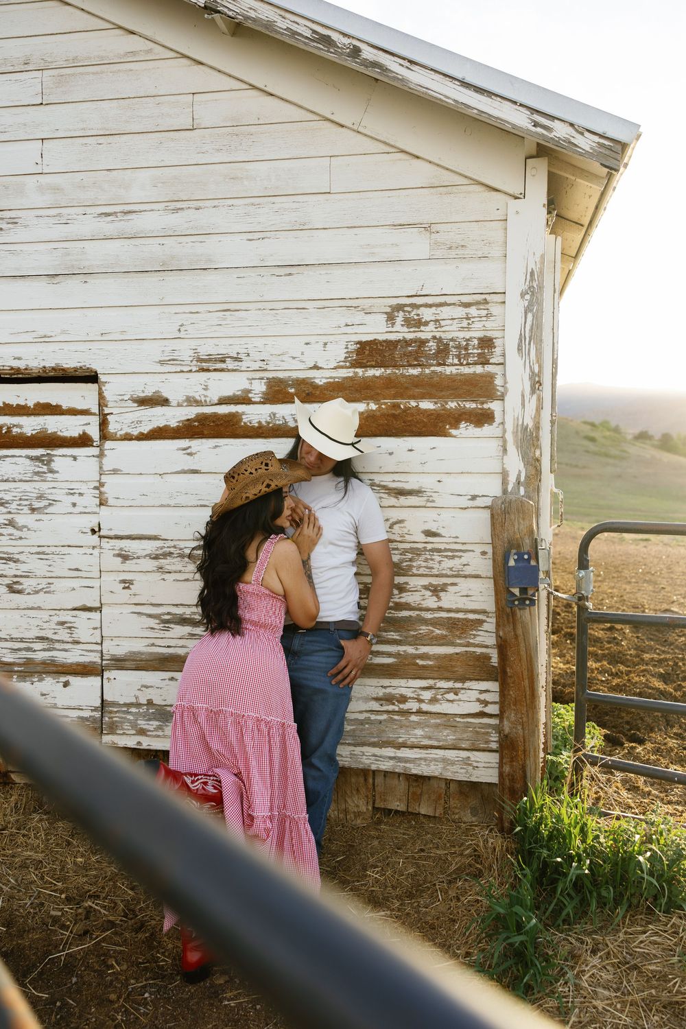 Couple in cowboy hats sharing a romantic moment leaning against a rustic white barn on a farm.