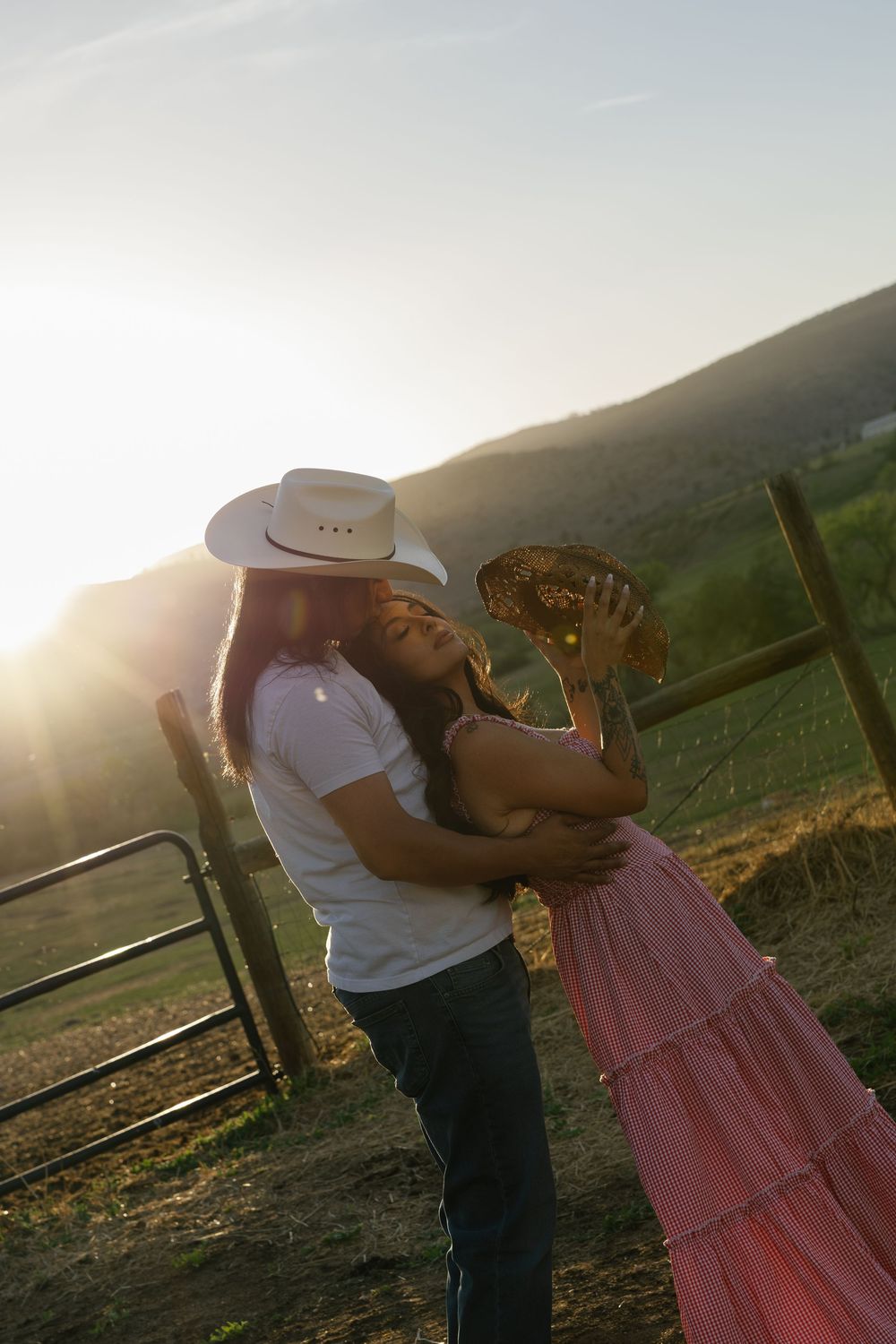 Couple in cowboy hats embracing at sunset by a ranch fence, woman in pink skirt leaning back romantically.